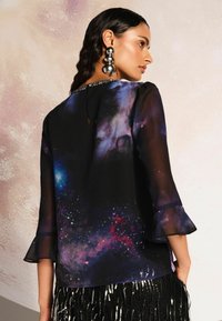 Áttetsző fekete blúz galaxis mintával, fodros ujjal és gyöngyökkel díszített nyakkivágással. A stílus lazán illeszkedik és hátul nyitott.