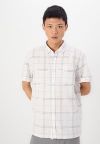 Chemise à manches courtes, col, de couleur blanche avec un tissu texturé, présentant un motif de grille noire et une poche poitrine unique.
