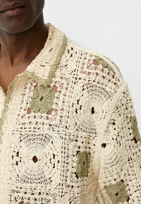 Camisa de crochet en crema y verde oliva, con patrones intrincados, cuello con solapa y cierre de botones con un acabado texturizado.