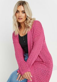 Lång, öppen stickad kappa i livlig rosa, med diamantmönster och ribbade muddar. Lager på en svart topp, parat med ljusblå jeans.