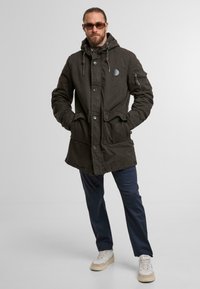 Dunkelolivgrüner Parka mit Kapuze, vorderer Knopfverschluss, zwei großen Vordertaschen und einem Patchdetail auf der linken Brust.