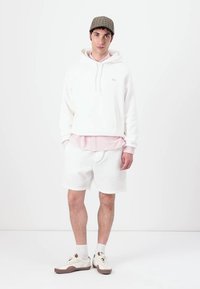 Felpa bianca con cappuccio e coulisse, indossata sopra una camicia rosa con colletto, abbinata a pantaloni corti bianchi testurizzati e sneakers crema. Cappello a scacchi marrone.