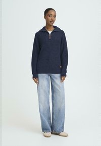 Navy gebreide trui met een ritskraag, voorzien van een gestructureerd patroon en ribbels bij de manchetten, gecombineerd met lichtblauwe wijde jeans en beige schoenen.