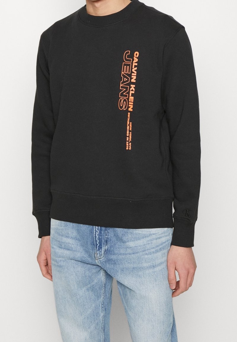 Sweat noir avec texte orange vertical "Calvin Klein Jeans" sur la poitrine, porté avec un jean en denim bleu clair.