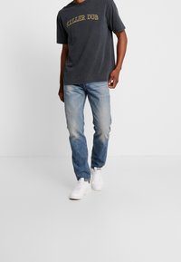 Tmavě šedé tričko s nápisem "KILLER DUB" žlutým písmem. Světle modré džíny, mírně vybledlé, a bílé tenisky. Jednoduchý design a volný střih.