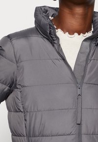 Chaqueta acolchada gris con un alto cuello, cierre de cremallera y detalles de costura horizontal. La tela parece suave con una textura ligera.