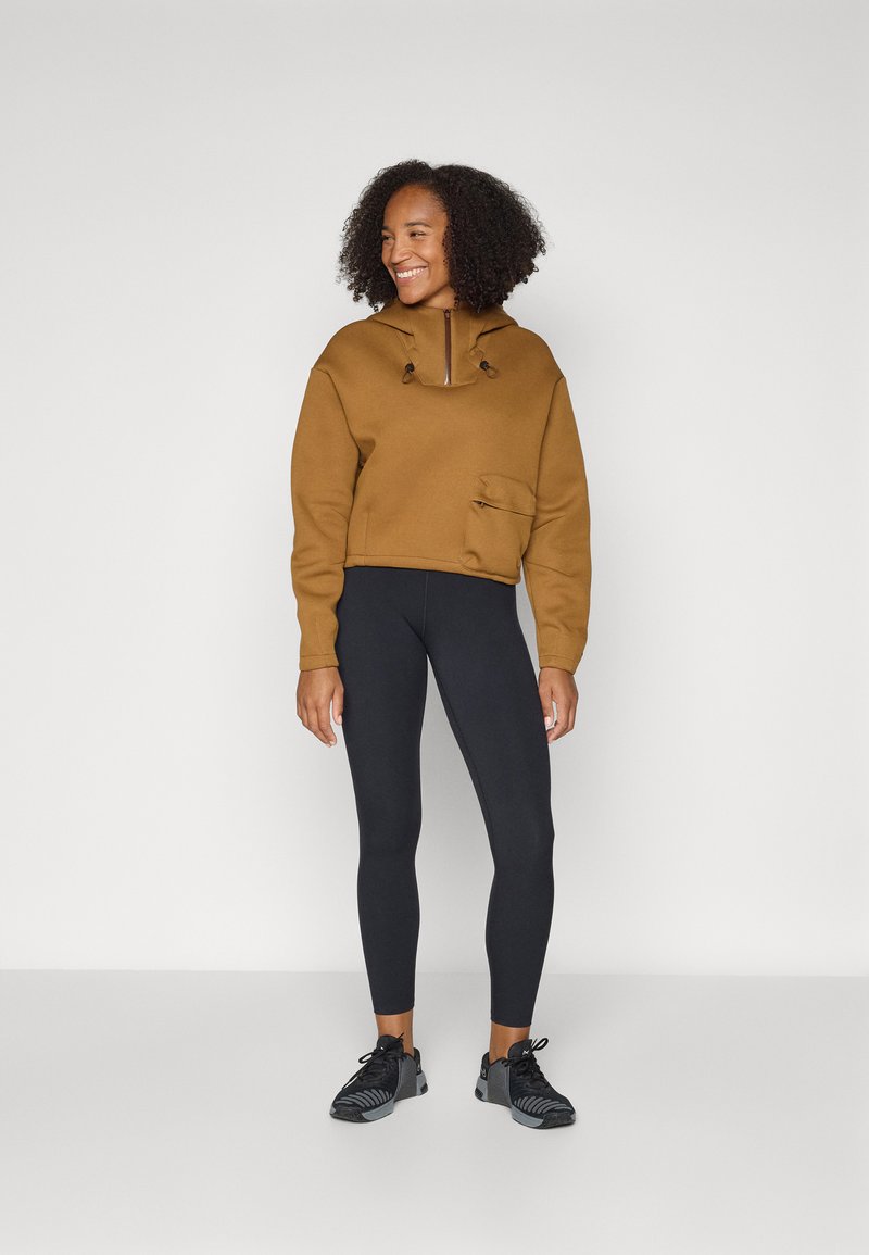 Eine lächelnde Frau trägt einen braunen Hoodie, schwarze Leggings und schwarze Sportschuhe und steht vor einem schlichten weißen Hintergrund.