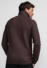 Sweater de punto marrón oscuro con cuello alto, dobladillo acanalado y costuras laterales. La textura parece suave y gruesa. Diseño liso sin patrones.
