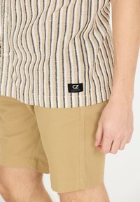 Randig beige skjorta med svarta linjer och en svart logopatch, kombinerad med kaki shorts. Skjortan har en knapp-för-ner design och en mjuk textur.