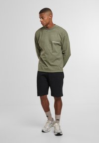 Vīrietis stāv ar rokām aiz muguras, ģērbies olīvzaļā garām piedurknēm kreklā, melnās šortos, bēšās zeķēs un baltās sporta kurpēs uz vienkārša fona.