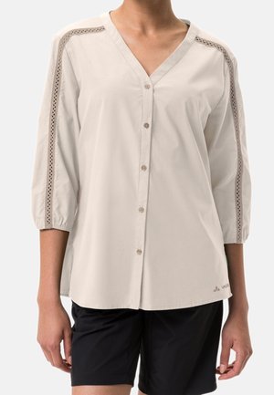 Vrouw draagt een beige V-hals blouse met knopen aan de voorkant en kantafwerking langs de mouwen, gecombineerd met zwarte shorts.