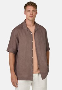 Camicia a maniche corte, con bottoni, di un marrone tenue, in misto lino con una finitura testurizzata; presenta un colletto e tasche laterali. Indossata sopra una maglietta rosa chiaro.
