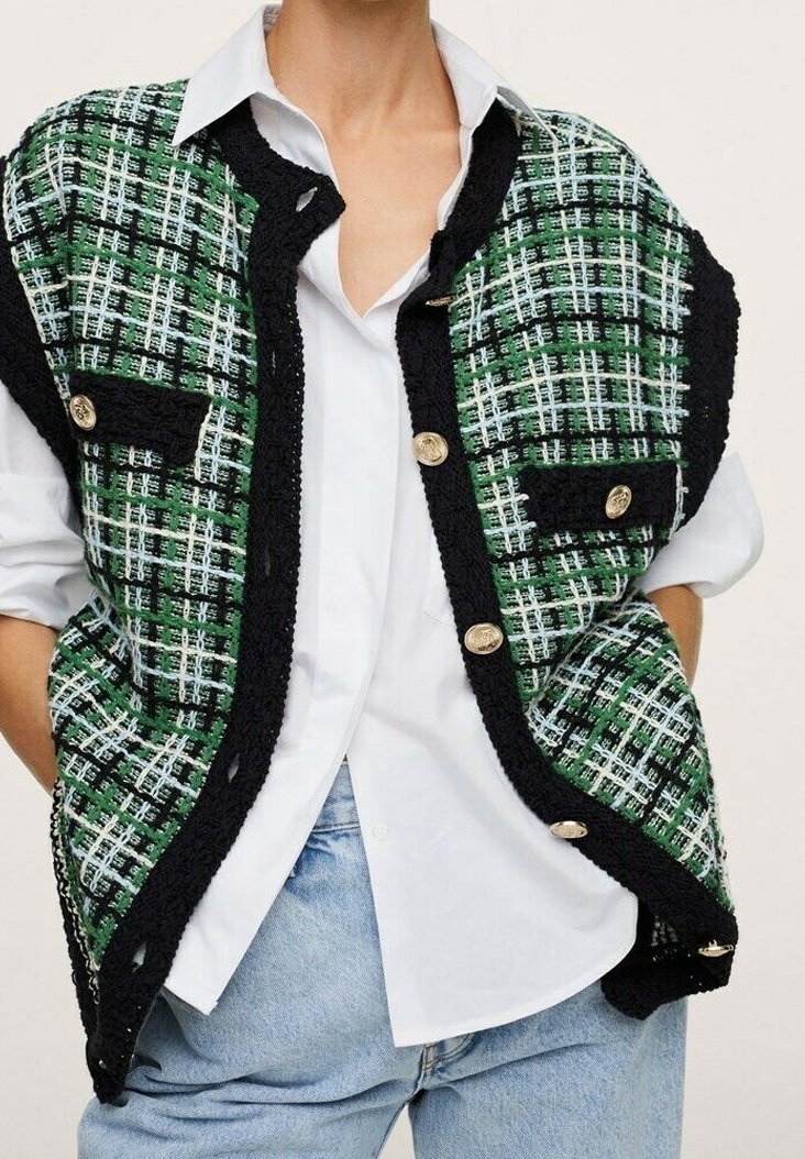Veste tricotée à carreaux verts, noirs et blancs avec des boutons dorés portée sur une chemise blanche à boutons et un jean bleu clair, les mains dans les poches.
