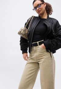 Νέα γυναίκα που φοράει μαύρο μπουφάν μπόμπερ, μαύρο τοπ, μπεζ παντελόνι με μαύρη ζώνη, κρατώντας τσάντα στον ώμο με σχέδιο από δέρμα φιδιού.