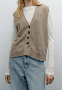 Beige neuleliivi, jossa on V-pääntie, napitettu etuosa, ribattu helma ja rento malli, kerroksittain valkoisen pitkähihasen paidan päällä.