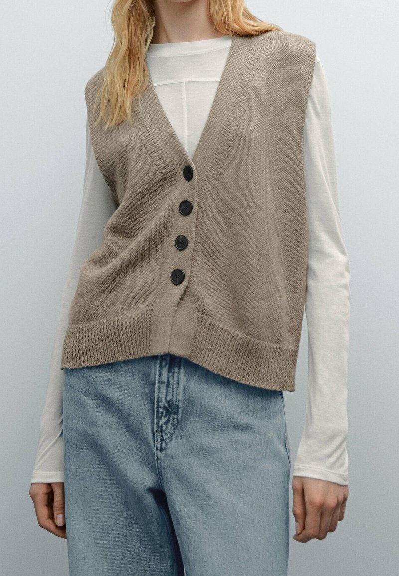 Beige neuleliivi, jossa on V-pääntie, napitettu etuosa, ribattu helma ja rento malli, kerroksittain valkoisen pitkähihasen paidan päällä.