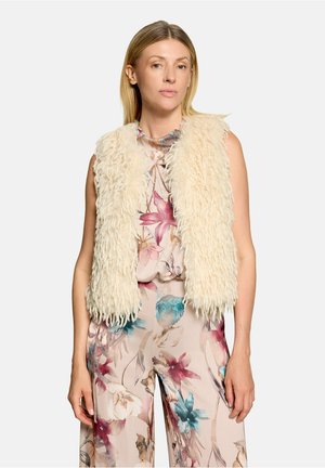 Vrouw draagt een crèmekleurige, fluffy bodywarmer over een mouwloze jumpsuit met roze, blauwe en beige bloemen en staat tegen een effen achtergrond.