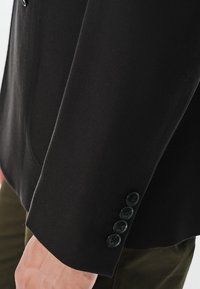 Czarny rękaw marynarki z teksturowanej tkaniny; posiada cztery czarne guziki i kołnierz z wycięciem, elegancko opadając na zielony strój.