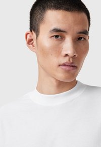 Weißes Baumwoll-T-Shirt mit einem hohen Rundhalsausschnitt, glatter Textur und minimalistischem Design, getragen von einer Person mit kurzen Haaren und dezenten Gesichtsmerkmalen.