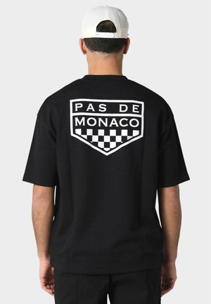 Camiseta de manga corta negra con un gran logo blanco "PAS DE MONACO" y un patrón de cuadros en la parte posterior, hecha de suave tela de algodón.