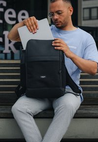 Hombre con gafas guarda un portátil en una mochila Larkson negra mientras está sentado en un banco de madera en el exterior.