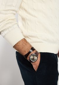Reloj de cuero marrón con herrajes plateados y una esfera gris claro. Suéter de punto en color crema con un patrón texturizado, combinado con pantalones de pana azul oscuro.