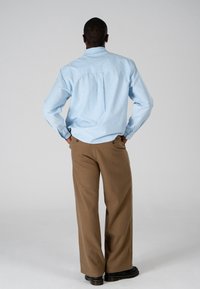 Homme debout, mains dans les poches, portant une chemise rayée bleu clair, un pantalon marron à jambes larges et des chaussures noires, tourné de dos sur un fond uni.