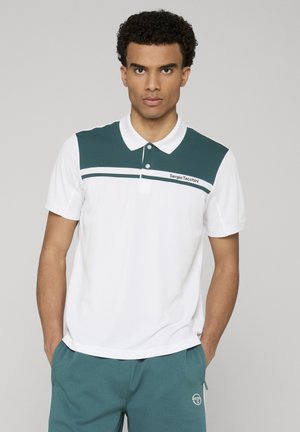 Uomo che indossa una polo bianca con pannello superiore verde scuro e logo, abbinata a pantaloncini verde scuro, mani nelle tasche, in piedi davanti a uno sfondo semplice.