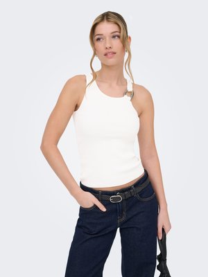 Vrouw met blond haar in een witte mouwloze top met ringdetail, donkere spijkerbroek, zwarte riem, die een zwarte tas vasthoudt tegen een effen achtergrond.