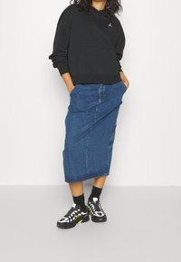 Sudadera negra con capucha y bolsillo frontal, combinada con una falda vaquera azul hasta la rodilla y zapatillas multicolores. Se ven calcetines oscuros con textura.