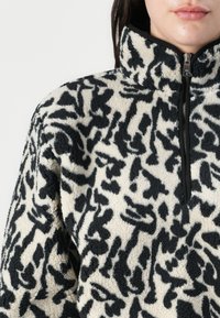 Pullover in pile color crema con una stampa leopardata astratta nera, caratterizzato da un colletto alto e una chiusura frontale con zip. Texture morbida con un fit rilassato.