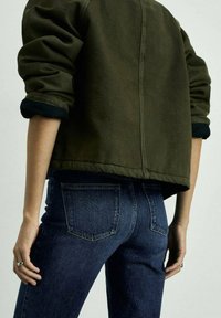 Olivgrön kort jacka med rullade svarta manschetter, i kombination med mörkblå denimjeans. Tyget verkar slätt med subtila sömnadsdetaljer.
