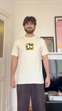 Άνδρας με σγουρά μαλλιά και γενειάδα, φορώντας λευκό μπλουζάκι με πράσινο και πορτοκαλί λογότυπο, στέκεται σε σαλόνι με έργα τέχνης και τηλεόραση πίσω του.