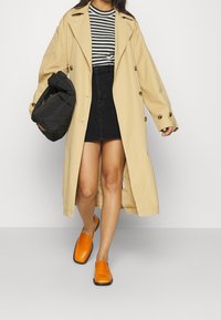 Trench-coat beige avec de gros boutons, jupe en denim noire, haut rayé noir et blanc, et chaussures orange sans lacets. Tenant un sac noir.