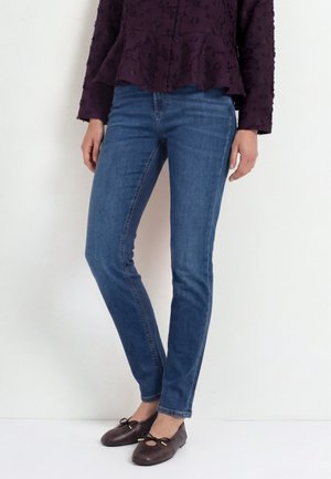 Frau trägt blaue Skinny Jeans, ein strukturiertes dunkelviolettes Oberteil und dunkelbraune Ballerinas mit Schleifendetails, steht vor weißem Hintergrund.