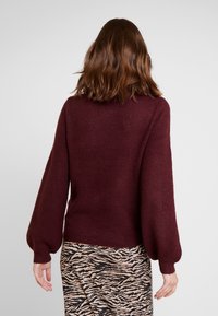 Pull en tricot bordeaux avec manches bouffantes et poignets côtelés, associé à une jupe imprimée zèbre. Le tissu semble doux et texturé.