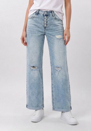 Osoba nosi svetlomodré vysoký pás široké nohavice džínsy s gombíkovým zapínaním, roztrhnutými kolenami, zrolovanými lemami, bielymi teniskami a vsadeným bielym tričkom.