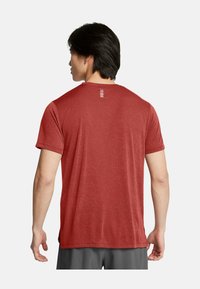 Camiseta atlética roja con un tejido texturizado, mangas cortas y un pequeño detalle de corte horizontal en la parte posterior. Pantalones cortos grises visibles por debajo.
