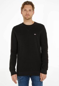 Zwart gebreid sweatervest met lange mouwen, ronde halslijn en geribbelde manchetten. Klein logo op de borst. Draag met lichtblauwe jeans.