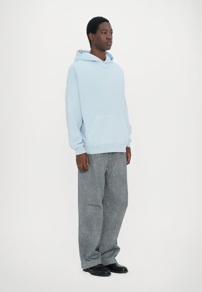 Felpa blu chiaro con tasca frontale e cappuccio con cordino, abbinata a pantaloni grigi a gamba larga, indossata con scarpe nere. Sfondo semplice.