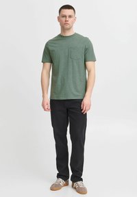 Camiseta verde con textura, de manga corta y con un bolsillo en el pecho, combinada con pantalones negros y zapatillas grises con rayas blancas.