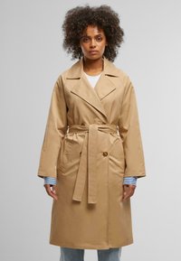 Beige trench coat with a double-breasted design, wide lapels, and a self-tie belt. Features long sleeves and a smooth, water-resistant fabric.