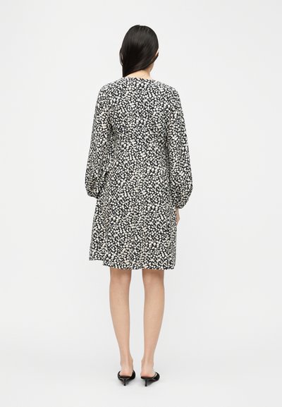 Γυναίκα με μακριά σκούρα μαλλιά, που φοράει μακρυμάνικο φόρεμα με μαύρα και λευκά λουλούδια και μαύρα παπούτσια με χαμηλό τακούνι, στέκεται γυρισμένη ανάποδα σε απλό φόντο.