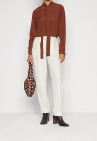 Blouse couleur rouille avec détail à nouer, pantalon crème, sac à poignée chaîne marron et bottines à talons noires. Tissu lisse et coupe structurée.