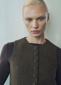 Brązowa sukienka w wzór houndstooth o dopasowanym kroju, okrągłym dekolcie oraz zapięciu na guziki z przodu. Czarna bluzka z długim rękawem pod spodem.