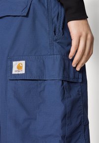 Siniset työhaalarit, jotka on valmistettu kestävästä kankaasta ja joissa on paikka-tasku, jossa on näkyvä keltavalkoinen logo. Kankaan teksturoidun pinnan yksityiskohdat ovat nähtävissä.