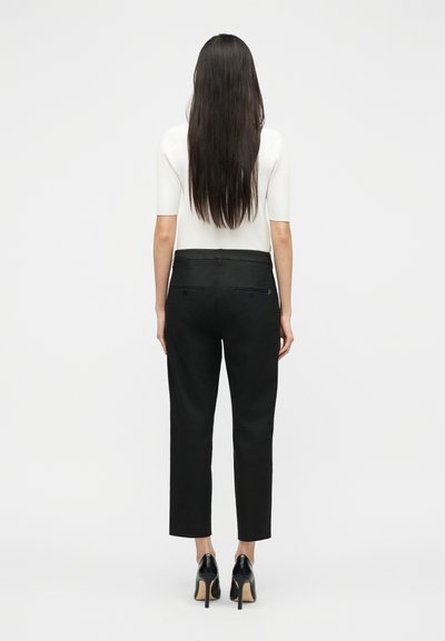 Γυναίκα με μακριά σκουρόχρωμα μαλλιά, φορώντας λευκό κοντομάνικο τοπ, μαύρο ψηλόμεσο παντελόνι και μαύρες ψηλοτάκουνες γόβες, στέκεται με την πλάτη προς το μέρος μας σε λευκό φόντο.