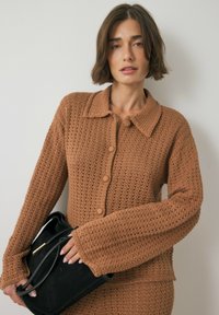 Brown knitted cardigan with a collared neck, button closure, and textured pattern. Model holds a black bag, showcasing the outfit's details.