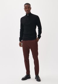 Suéter negro con textura y cuello con cremallera, combinado con pantalones ajustados marrones y zapatos negros sin cordones. El conjunto es minimalista y contemporáneo.