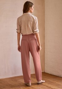 Gestreiftes beiges Hemd mit aufgerollten Ärmeln, kombiniert mit hoch geschnittenen rosa Hosen mit Gesäßtaschen. Hintergrund aus Holzfußboden.